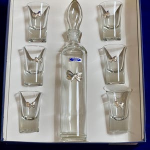 May include: A clear glass drinks set comprising a decanter and six shot glasses, each decorated with a silver bow. The decanter is tall and slender with a glass stopper. The set is presented in a white box with a blue border.