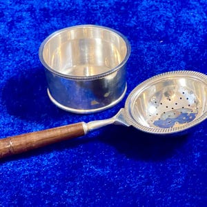May include: A silver tea strainer with a wooden handle and a matching silver bowl. The strainer has a perforated bowl shape. The wooden handle is brown. The bowl is round and silver. The items are on a blue surface.