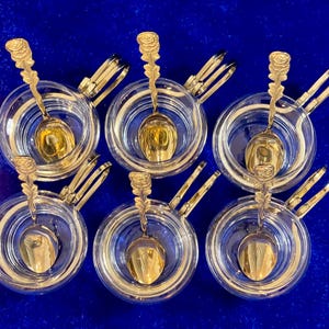 Könnte beinhalten: Sechs klare Glasschalen mit goldfarbenen Löffeln und Zangen, vor einem tiefblauen Hintergrund. Die Löffel haben ein Rosendesign am Griff. Die Zangen sind an den Schalen befestigt.