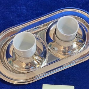 Può includere: Un vassoio d'argento con due tazze in ceramica bianca. Le tazze hanno piccole maniglie e poggiano su piattini abbinati. Il vassoio è di forma ovale e riflette la luce. Il set è su una superficie blu.