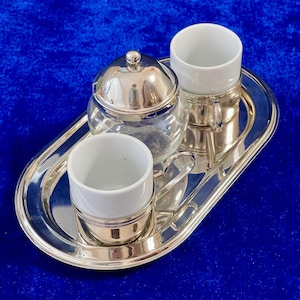 Op de afbeelding: Een zilveren koffieset op een zilveren dienblad. De set bevat twee witte keramische kopjes, een glazen suikerpot met een zilveren deksel en een zilveren dienblad. De set wordt gepresenteerd op een blauw oppervlak.
