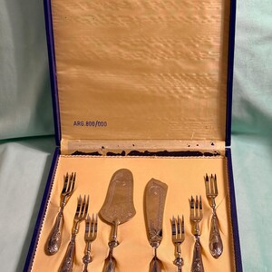 Puede incluir: Un juego de cubiertos plateados en una caja de presentación azul con interior color crema. El set incluye seis tenedores, un servidor de pasteles y un cuchillo, todos con mangos grabados. El texto ARG.800/000 está impreso en el interior.