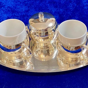 Puede incluir: Un juego de café plateado sobre una bandeja plateada. El juego incluye dos tazas de porcelana blanca con detalles plateados, un azucarero con tapa y una bandeja de servicio. El juego se exhibe sobre una superficie azul.