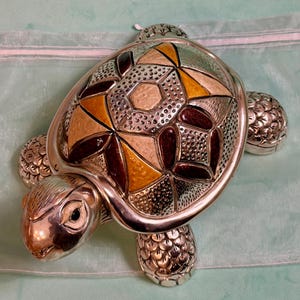 Op de afbeelding: Een zilverkleurig metalen schildpadbeeldje met een decoratief schild met geometrische patronen in tinten amber, donkerbruin en getextureerd zilver. De schildpad heeft een gepolijste metalen afwerking, gedetailleerde schubben en een gladde kop.