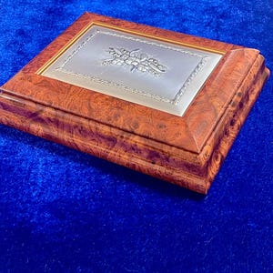 Puede incluir: Una caja de joyería rectangular de madera con una tapa plateada. La caja tiene un acabado de veta de madera marrón rojizo y un diseño floral plateado en la tapa. La caja está sobre un fondo azul.