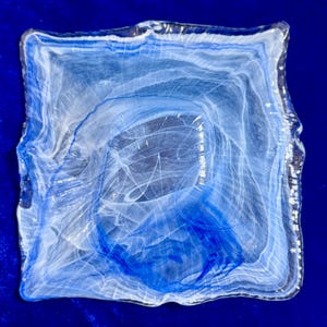 Op de afbeelding: Een vierkante glazen schaal met een wervelend patroon van doorschijnende witte en diepblauwe kleuren. Het glas heeft een golvende, onregelmatige rand en een glad, gepolijst oppervlak, wat een gemarmerd effect creëert in de gehele vierkante schaal.