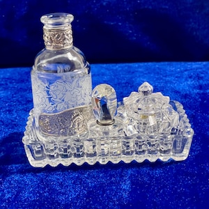 May include: A clear glass vanity set with a silver-colored metal accent. The set includes a perfume bottle, a small jar with a lid, and a crystal-like applicator, all resting on a rectangular tray. The bottle has a floral design.