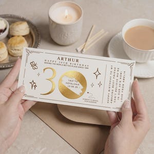 May include: A cream-coloured birthday scratch card with gold foil details. The card says "ARTHUR HAPPY 30TH BIRTHDAY" and features a scratch-off area for a trip to Kefalonia. The card is held by a person.