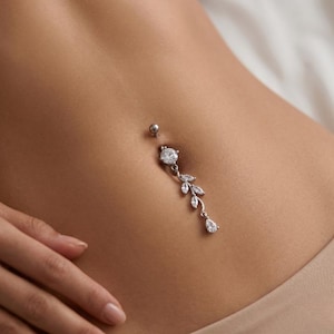 Può includere: Un piercing all'ombelico argentato con un design a foglie pendenti. L'anello presenta una grande gemma rotonda e trasparente nella parte superiore e una gemma a forma di goccia nella parte inferiore. Piccole gemme trasparenti sono incastonate nel design della foglia.