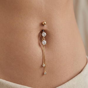 May include: A gold-coloured belly button ring with a dangling chain. The ring features three clear, round gemstones and a small ball on top. The jewellery is set against a light skin tone.