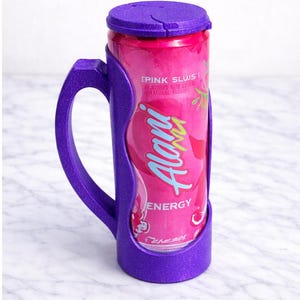 Op de afbeelding: Een paarse blikjeshouder met een handvat, met een roze Alani Energy-blikje. Het blikje heeft de tekst "Pink Slush" en "Energy". De houder heeft een bijpassende paarse deksel.
