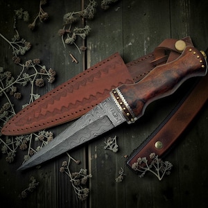 May include: A handcrafted dagger with a patterned steel blade and a dark wooden handle. The handle is adorned with brass rivets and a decorative band. The dagger rests in a brown leather sheath with embossed detailing, alongside a matching belt.