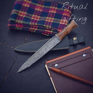 May include: A Damascus steel knife with a wooden handle and leather sheath. The knife is next to a brown leather notebook and a plaid fabric. The text "Ritual gifting" is in the upper right corner.