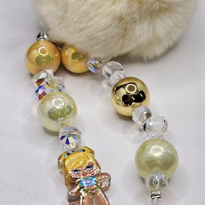 May include: A decorative keyring featuring a fluffy cream-coloured faux fur pom-pom attached to two beaded strands. The strands include iridescent peach beads, clear faceted crystals, a shiny gold sphere, and a character charm with blonde hair.