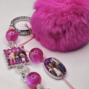 May include: A keychain featuring a large, vibrant hot pink faux fur pom-pom, a silver-toned metal ring with embedded crystals, and a pink cord strung with translucent pink beads, clear faceted beads, and two charms depicting groups of fashion dolls.