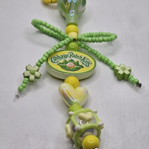 May include: A handmade keychain featuring a green and yellow design with a Cabbage Patch Kids logo. The keychain includes a yellow loop, green beads, and decorative elements, including a heart and flower shapes. The text "Deezbeadz" is visible.