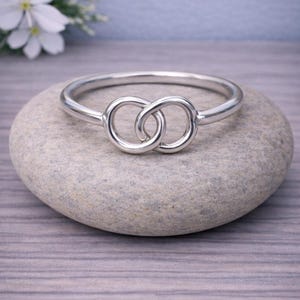 Könnte beinhalten: Ein silberfarbenes Armreif-Armband mit einem Unendlichkeitsknoten-Design. Das Armband liegt auf einem glatten, grauen Stein. Das Design zeigt zwei ineinandergreifende Kreise. Weiße Blumen im Hintergrund.