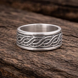 Puede incluir: Un anillo plateado con un detallado diseño trenzado alrededor de la banda. El anillo se muestra sobre una superficie de madera rústica, mostrando su intrincado diseño y brillo metálico. El diseño del anillo es un patrón continuo e entrelazado.