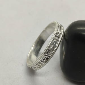 May include: A silver ring with an intricate, repeating pattern. The ring is leaning against a smooth, black, rounded stone. The ring's design features detailed floral and geometric elements, creating a textured surface. The ring is likely made of sterling silver.