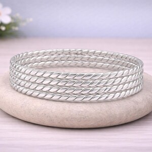 May include: A stack of four silver-colored, twisted rope-style bangles rests on a light-colored, oval stone. The bangles are made of a shiny metal. The background includes a blurred floral arrangement and a light-colored surface.