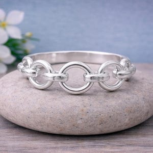 May include: A silver-colored bangle bracelet with a chain-link design. The bracelet is composed of interconnected circular links. It rests on a smooth, gray stone, with white flowers and a blue background.