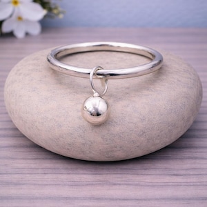 Könnte beinhalten: Ein silbernes Armreif-Armband mit einem hängenden Kugelanhänger. Das Armband ist rund und glatt, mit einem kleinen, runden Anhänger, der an einer Schlaufe hängt. Der Schmuck wird auf einem hellgrauen Stein präsentiert, mit weißen Blumen im Hintergrund.