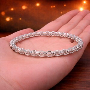 Könnte beinhalten: Ein silbernes Armband mit einem Kettenglied-Design. Das Armband ist kreisförmig und scheint aus einem glänzenden, metallischen Material zu bestehen. Das Armband liegt auf der offenen Handfläche einer Person, mit einem verschwommenen Hintergrund.