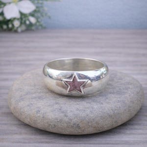 May include: A silver ring with a star design. The ring has a wide band and a raised star in the center. The star is a muted pink color. The ring is sitting on a gray stone.