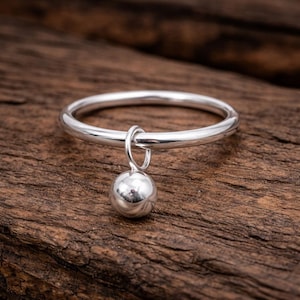 Puede incluir: Un anillo de plata con un pequeño dije de bola colgante. El anillo está sobre un fondo de madera rústica. El dije es una pequeña esfera brillante, que añade un toque de elegancia a la banda simple. El anillo es una joya.