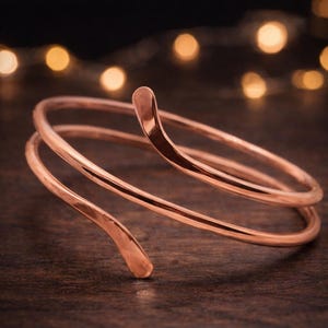 May include: A copper-colored arm cuff with a spiral design. The cuff wraps around the arm with a smooth, polished finish. The ends of the cuff have a curved, hook-like shape. The background is blurred with warm, golden lights.