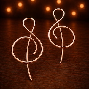 May include: A pair of rose gold-colored metal earrings shaped like treble clef musical symbols. The earrings feature a continuous, smooth wire design that forms the iconic G-clef shape, with a polished, reflective surface that catches the light.