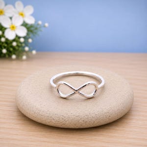May include: A polished silver ring featuring a central infinity symbol design. The band is smooth and circular, showcasing a continuous metallic finish that forms the horizontal figure-eight shape. The ring is displayed in a clear, focused view.