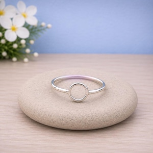 Puede incluir: Un anillo fino de tono plateado pulido que presenta un diseño circular calado en el centro. La banda es lisa y de grosor uniforme, creando un bucle metálico continuo que se conecta con el elemento circular central en ambos lados.