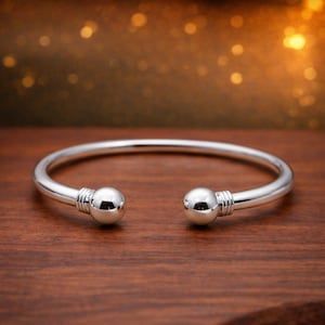 May include: A polished silver cuff bracelet featuring an open design with two spherical ball ends. Each end is accented with three subtle horizontal grooves near the base of the sphere, creating a clean and reflective metallic finish.
