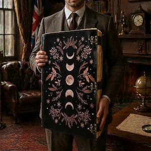 May include: A large black leather-bound book featuring a vertical sequence of moon phases in copper foil, surrounded by intricate botanical illustrations of leaves and snowflakes. The book is secured with three antique brass metal clasps.