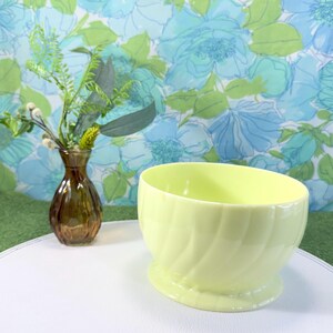 May include: A pale yellow ceramic bowl with a spiral design, set on a small base. A small brown glass vase with greenery is to the left. The background features a floral pattern in shades of blue and green.