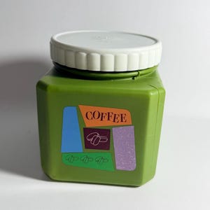 May include: Green plastic coffee container with a white lid. The container has a rectangular shape with a colorful label that says "COFFEE" and features coffee bean graphics. The container is likely for storing coffee.