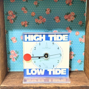 May include: A square tide clock with a light blue circular face featuring numbers one through five, a sailboat icon, and a red circular indicator. The clock has black hands and the text HIGH TIDE and LOW TIDE printed in bold white letters.
