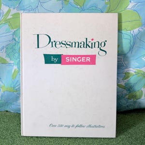 May include: A white hardcover book titled Dressmaking by SINGER with dark green and pink text on the front cover. The cover includes the subtitle Over 350 easy-to-follow illustrations printed in a small dark green font at the bottom center.