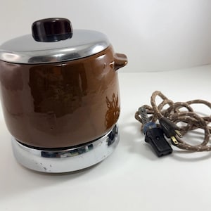 May include: A vintage brown electric fondue pot with a silver lid and base. The pot has two handles and a dark brown knob on top. A brown and white cord with a plug is next to the pot.