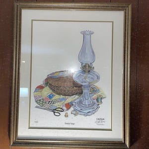 May include: Framed print of a still life featuring a glass oil lamp, a basket, a colorful quilt, and sewing tools. The lamp is clear with a brass base. The quilt has a patchwork design. The print is in a gold frame.