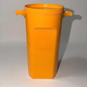 May include: A bright orange, rectangular pitcher with a textured rim and two small handles. The pitcher has a smooth, glossy finish and a slightly curved base. The design is simple and modern, suitable for serving beverages.