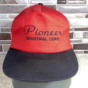May include: A red and black baseball cap with the text "Pioneer Industrial Corp." embroidered in black. The cap has a structured design with a curved brim and a black top button.