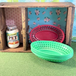 May include: A wooden crate with patterned paper lining, containing a glass jar of Instant Pet Nonfat Dry Milk, one red oval plastic serving basket, and a stack of three green oval plastic serving baskets, all arranged on a green surface.