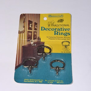 May include: A package of Homecraft brand decorative rings for hanging pictures, mirrors, plaques, and decoupage. The package has a yellow and blue color scheme and includes three metal rings with screws. The text on the package reads "8 Traditional Decorative Rings."