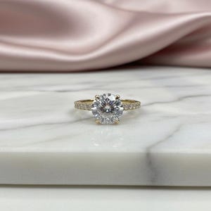 May include: A gold ring featuring a large, clear, round-cut gemstone. The gold band is embellished with smaller, clear gemstones. The ring is displayed on a white marble surface, with a pink satin fabric in the background.
