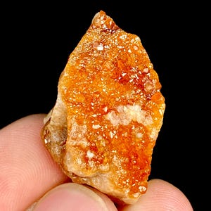 May include: A vibrant orange-hued crystal cluster with a rough, textured surface. The mineral displays a mix of crystalline structures and a warm, earthy color palette. The crystal is held between fingers against a black background.