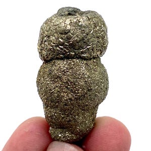 May include: A pyrite specimen, also known as fool's gold, with a layered, textured appearance. The mineral has a metallic, brassy-yellow color and is held in a hand against a white background. The pyrite is approximately 2 inches tall.