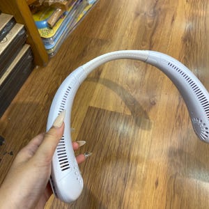 May include: A white, hands-free, wearable neck fan. The fan has a curved design that rests on the shoulders and neck. It features a series of vents and a flexible neckband. The fan is designed to provide personal cooling.