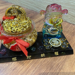 Könnte beinhalten: Eine goldfarbene Münze und ein Glasgefäß gefüllt mit goldfarbenen Objekten, gebunden mit einem roten Band und Quaste. Eine farbenfrohe Glasfigur steht auf einem schwarzen Sockel mit chinesischen Schriftzeichen.